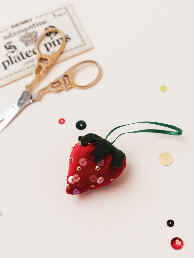 A velvet strawberry ornament on a white table with a scattering of sequins around