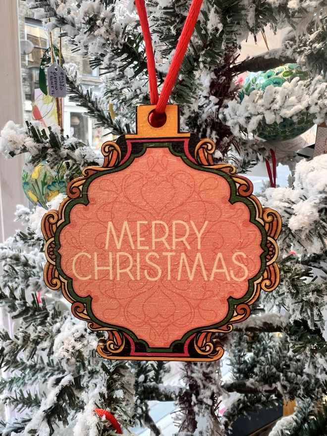 Multi coloured wooden tree decoration with 'Merry Christmas' written at the centre.  It is hanging by a red ribbon on a fake white christmas tree with other decorations. 