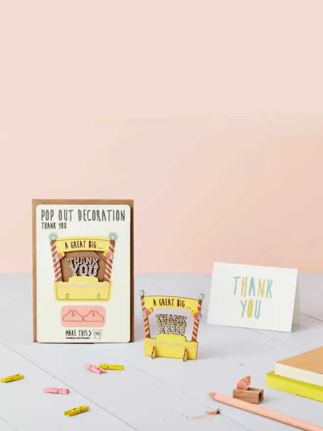 3D laser-cut thank you pop out decoration and thank you card and brown kraft envelope on top of a wooden desk in front of a peachy pink coloured background
