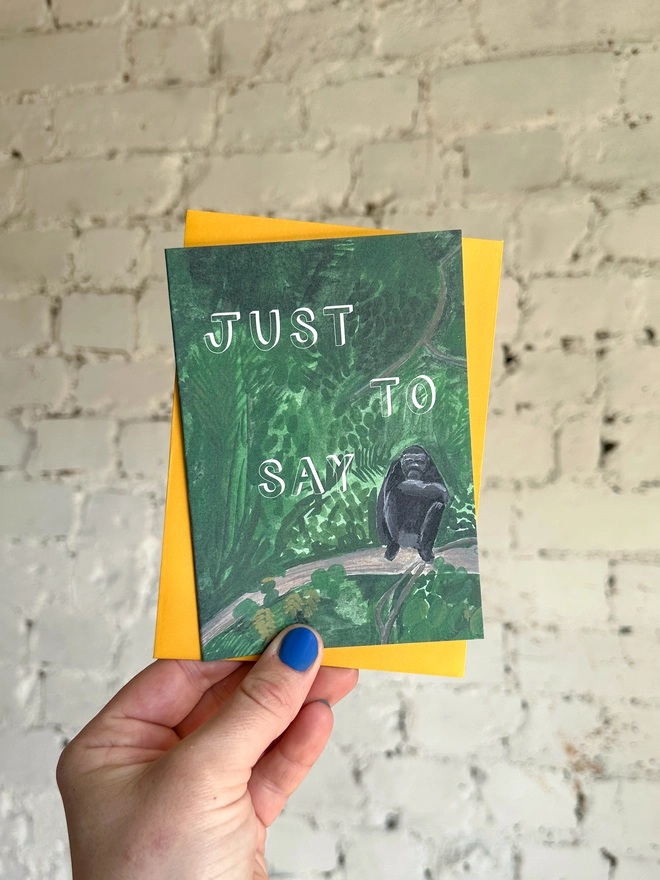 a greetings card featuring an illustration of a chimp sitting in the jungle with the text “just to say” with a deep yellow envelope