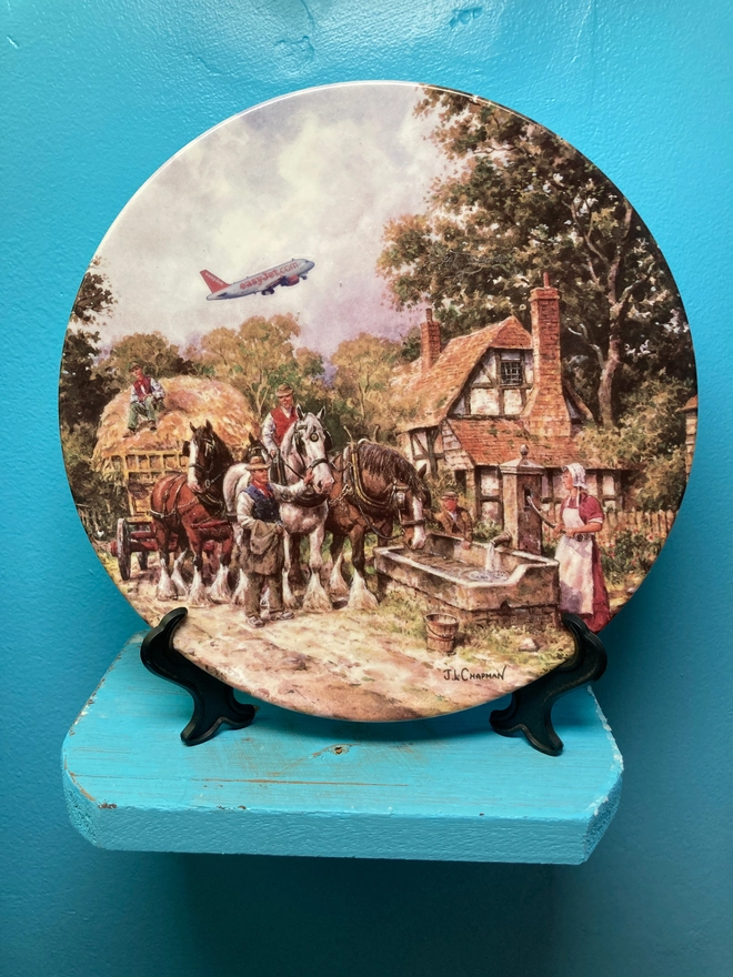 Decorative 8” plate featuring a chocolate box-twee cottage and a hay cart pulled by shirehorses. An easyJet plane soars off in the distance, causing a jarring contrast to the 17th century rural scene. 