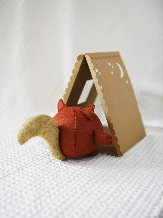 A handmade felt squirrel stands on a white surface beside a handmade cardboard house.