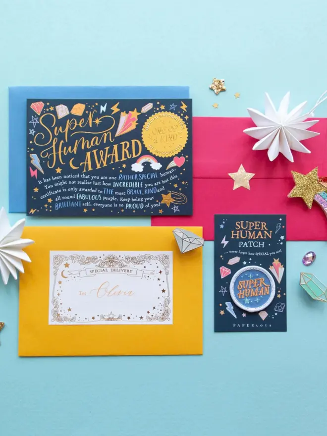 The complete set: award certificate, patch card, and a yellow envelope addressed to Olivia. Blue and pink envelopes, enamel gem pins and glitter stars are arranged around it.