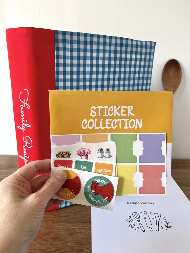 Close-up of hand holding yellow 'Sticker Collection' envelope and colourful index stickers, small square food pictures and phrases stickers and round stickers with hens and phrases. Blue gingham recipe binder with 'Family Recipes' embroidered in white thread on red spine, standing on wooden bench in background with 'Recipe Planner' page flat on bench.