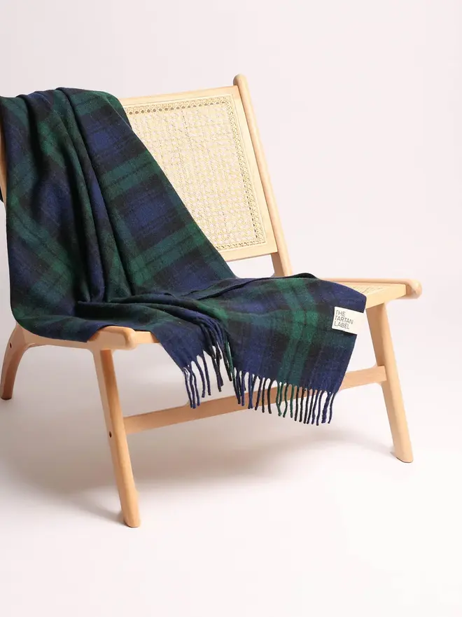 Black Watch Tartan Blanket, a green blanket draped over a wooden chair on a plain surface against a white backdrop. 