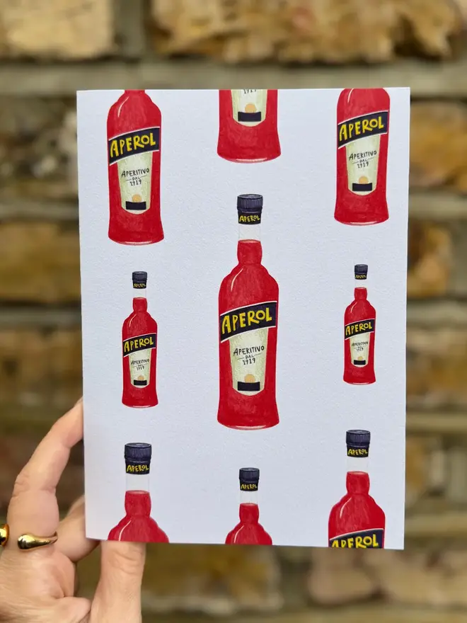 aperol spritz bottle illustrated card. card held against brick wall backdrop.