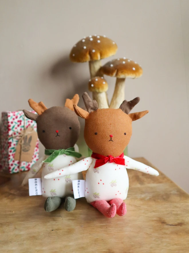 Two handmade reindeer dolls on a green background. Each doll has soft brown antlers, embroidered facial features, and wears a festive white outfit with a pinecone print. One doll has red legs and a red velvet bow; the other has green legs, a green bow, and the name “Arya” embroidered on its body. Both have a "BOKUNO" fabric tag.