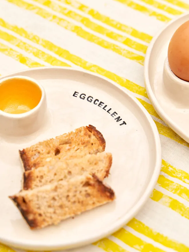 Egg And Soldier Plates, a plate with an integrated egg cup and toast soliders placed on a yellow striped surface. The wording reads 'eggcellent' 