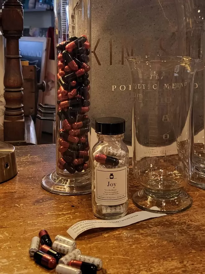 Glass bottle containing black and rose gold Joy poetry pills printed on banana paper.  Displayed on Poetry Pharmacy counter with medicine glass