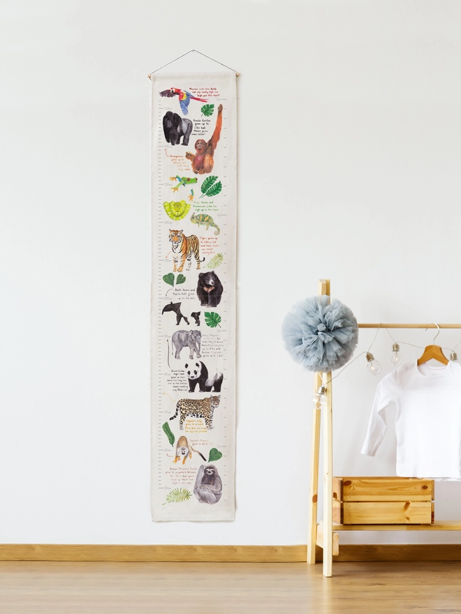image features the height chart hung on a wall next to some children’s furniture. The height chart has an off white background and features illustrations of animals and plants from jungle habitats all the way up it.