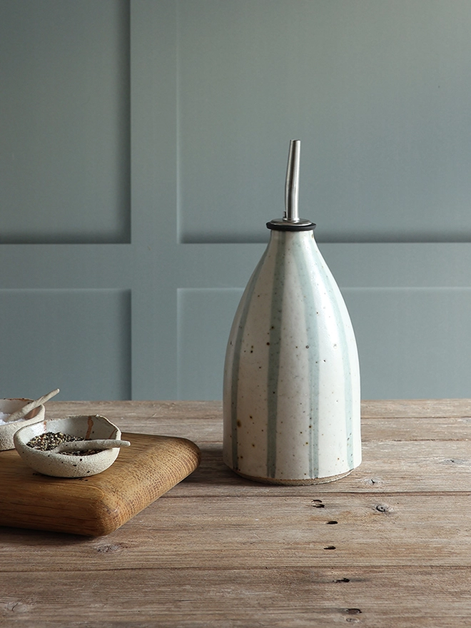 Handmade Ceramic Stoneware Oil Pourer in Blue and White Stripe sat on a wooden surface next to a wooden chopping board holding small handmade ceramic dishes containing salt and pepper. 