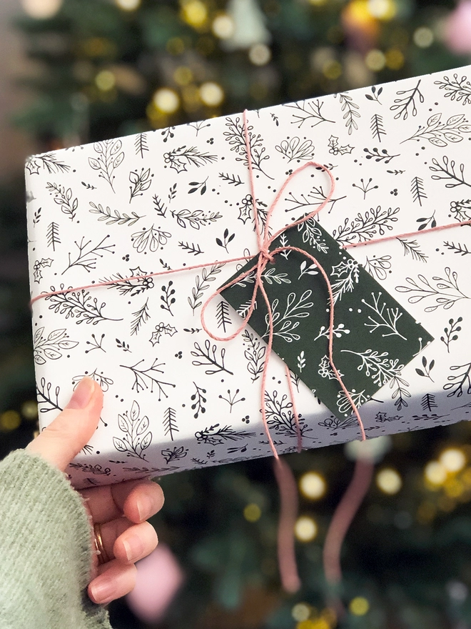 A gift wrapped in white wrapping paper with a Christmas botanical design is being held in front of a Christmas tree.