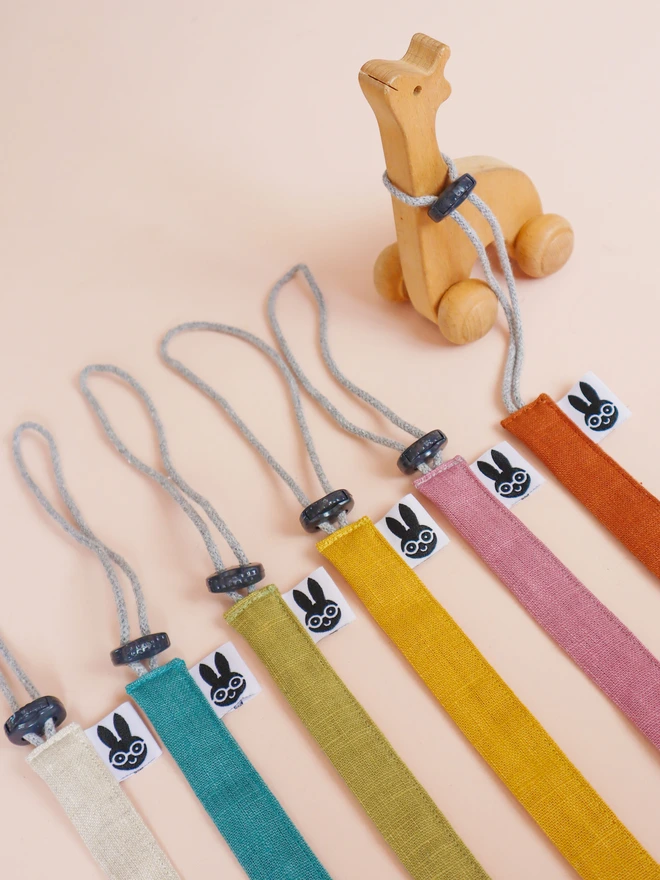 Handmade linen toy strap with grey string and a toggle attached to wooden baby toys, along with other colour options in rainbow colours.