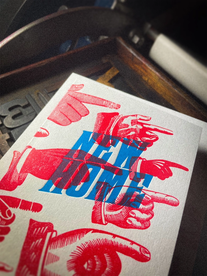 A double-sided, hand-printed letterpress artisan moving house announcement card with bold blue “New Home” typography and fluorescent red pointing hand illustrations (fists) on thick premium white card.