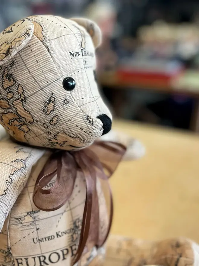 Teddy bear made from map-print fabric sitting upright on a wooden table, wearing a brown bow.