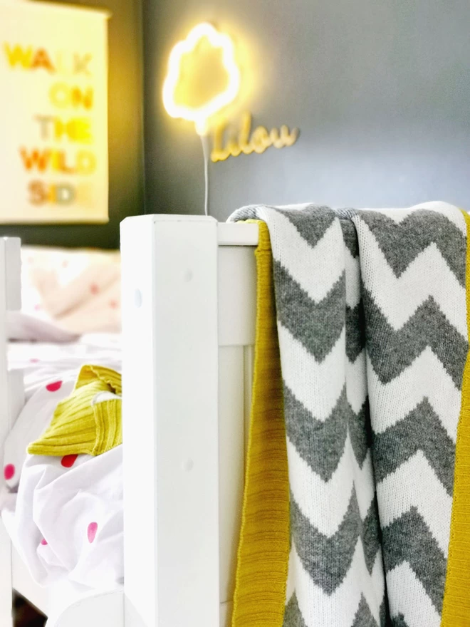 Close up of the end of a white single bed with a grey and white chevron blanket with mustard yellow trim draped over the end of the bed. On the wall is a child’s name plaque and a neon cloud wall light which is turned on creating a cosy bedtime mood.