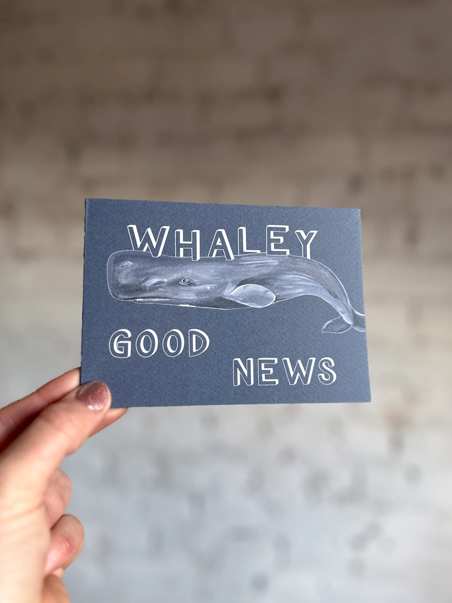 a dark grey blue greetings card featuring an illustration of a sperm whale with the text “whaley good news”