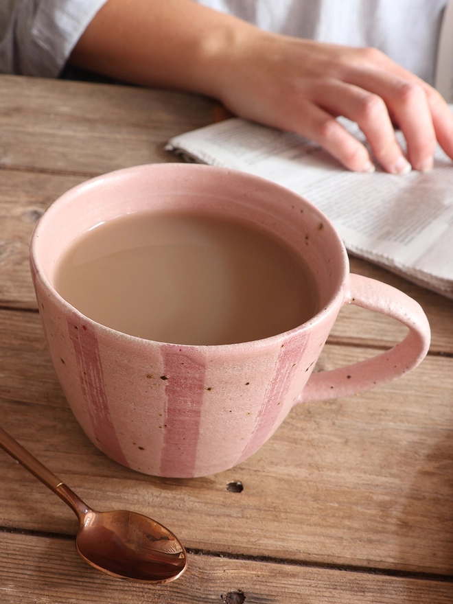 Handmade Ceramic Stoneware Mug in Pink Stripe sat on a wooden surface next to a brass spoon. A female hand on top of a newspaper can be seen in the background. 