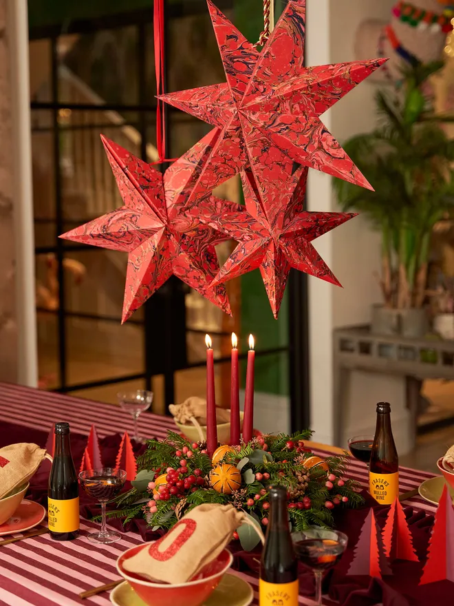 Red marbled paper hanging star decorations in various sizes (small, medium & large), hung above a busy Christmas tablescape from red ribbon