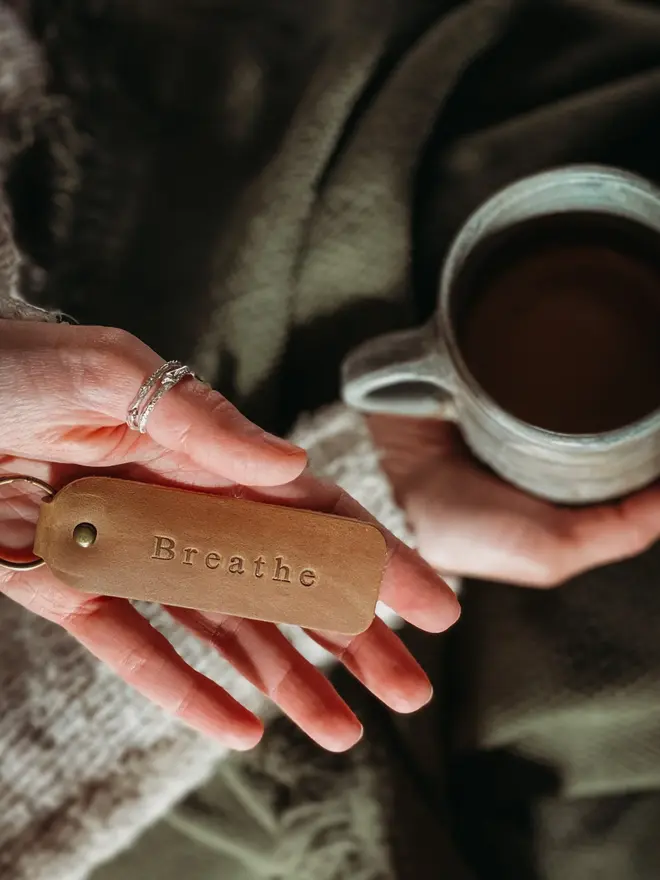encouraging leather keyring. a hand held brown leather keyring embossed with 'breathe' with a cup of coffee in the background.