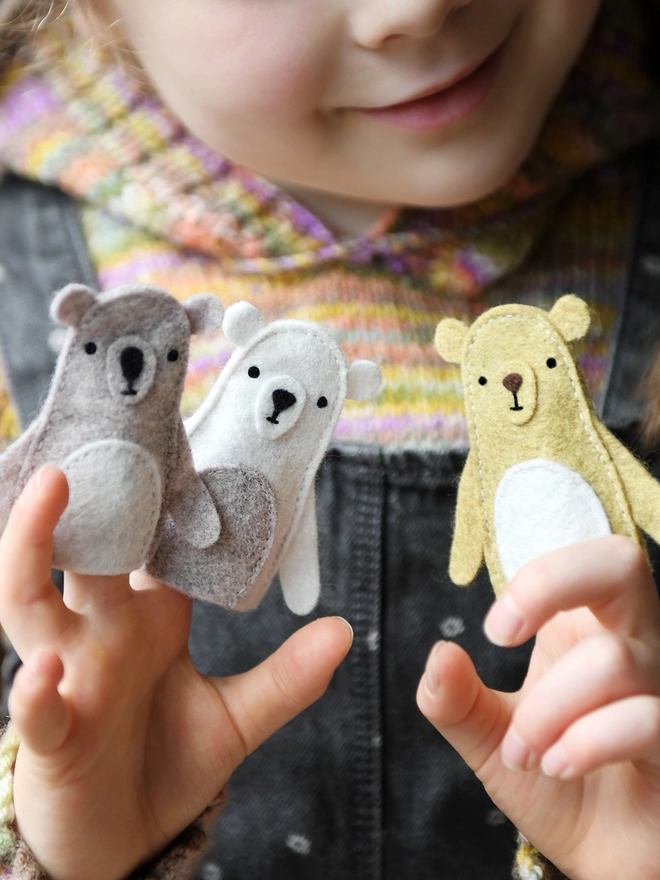 A young child wearing a knitted jumper and grey dungarees holds three handmade felt bear finger puppets on their fingers.