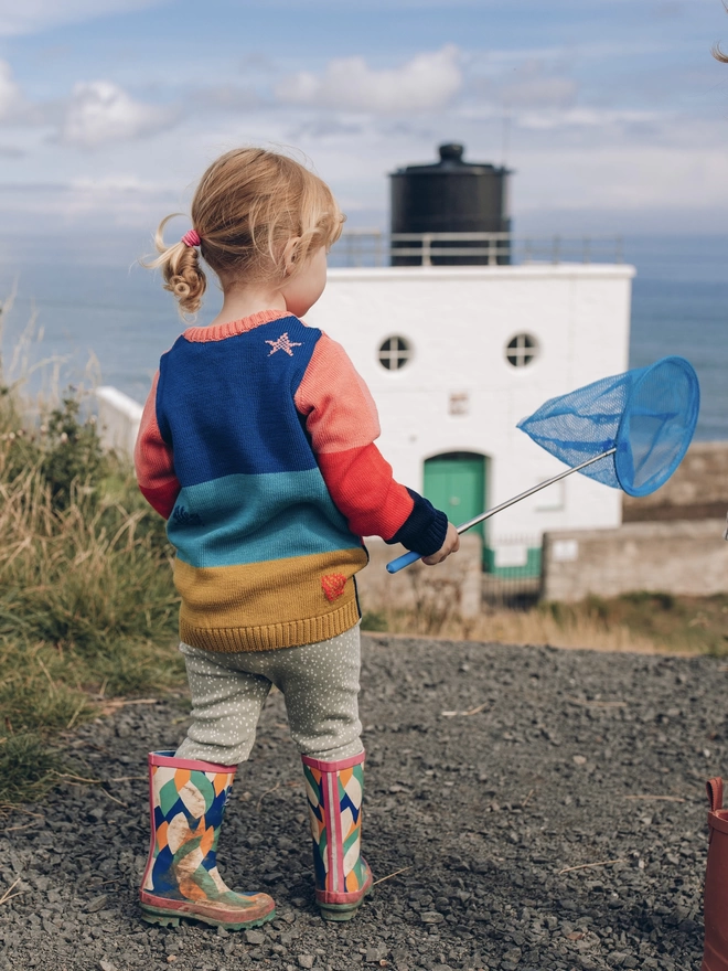 A little girl holding a blue fishing net near a lighthouse, dressed in 'The Stargazer' Knitted Jumper from The Faraway Gang.