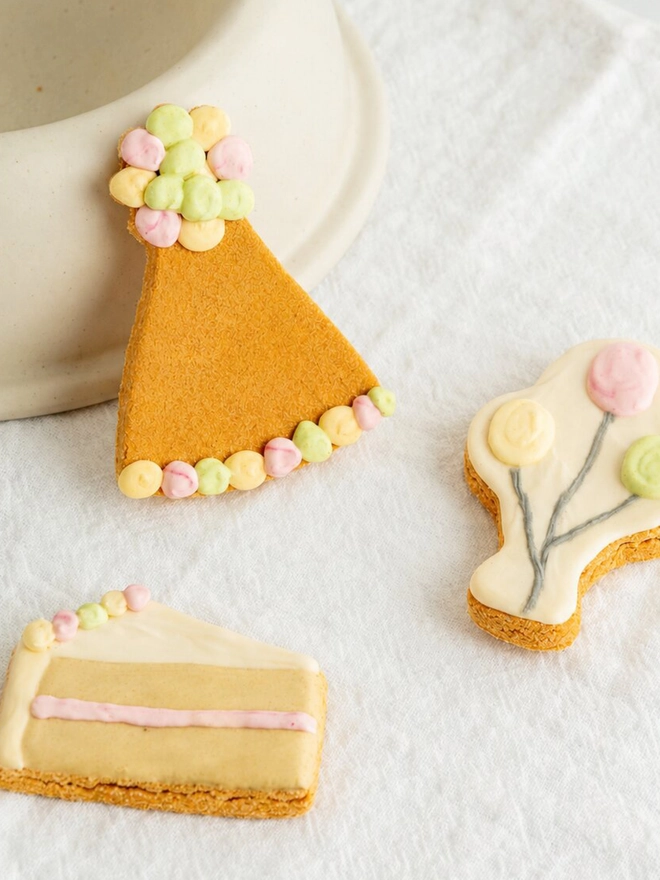 Celebration iced dog biscuit trio styled on white fabric beside a ceramic stand.