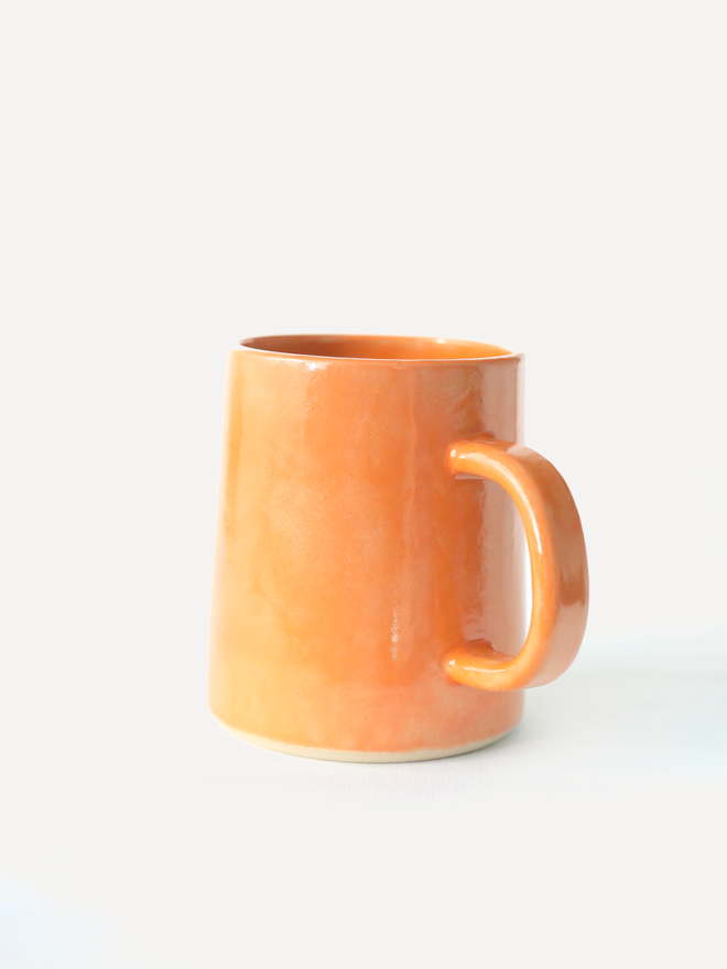 A handmade ceramic pint mug in mandarin orange glaze with a chunky loop handle, photographed against a plain light grey background