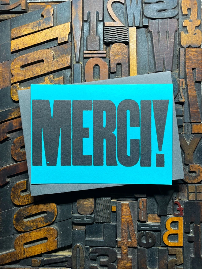 A bright turquoise thick duplex card with the word "MERCI!" printed in bold, black letters lies on a surface covered with various letterpress blocks.