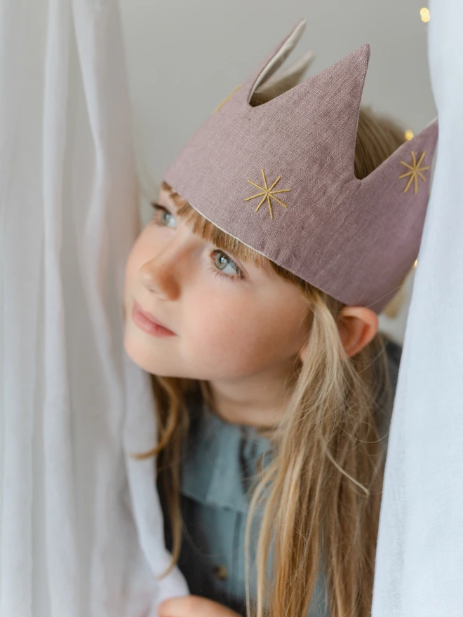 Girl in lilac linen birthday celebration crown peeping through the curtains