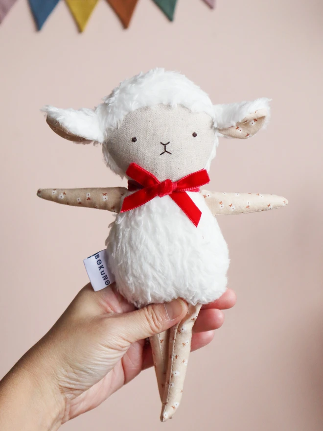 Handmade lamb doll with a fluffy white body, stitched face, beige floral fabric arms and legs, and a small red velvet bow tied around its neck.