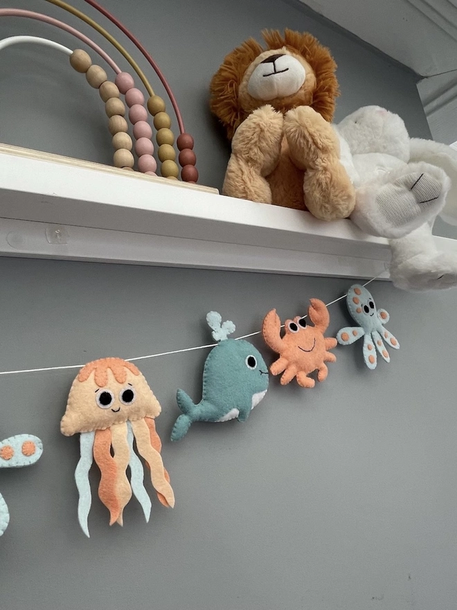Nursery Under The Sea Garland, a sea themed garland hanging from a white shelf against a grey backdrop in a child's bedroom. 