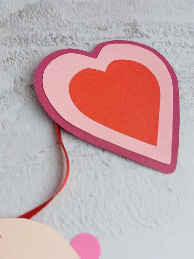A closeup of the garland end. A rapseberry pink heart, with a smaller baby pink heart layered on top, followed by a smaller bright red heart in the centre.