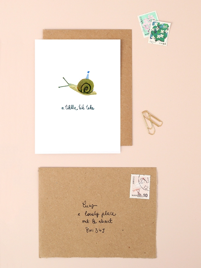 An image of a mini, rectangular white greetings card with a cute illustration of a snail with a tiny party hat on it's shell. A hand written 'a little but late' message in black ink is seen under the central illustration. The card is seen on top of a brown rectangular kraft envelope.