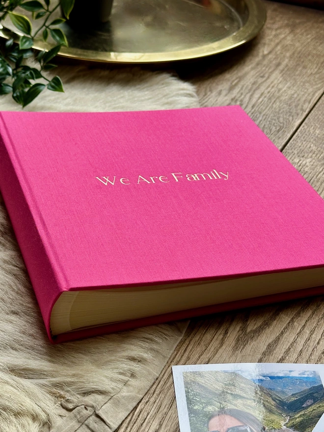 a pink photo album lies on a sheepskin rug and there is a gold tray in the background. The photo album has been printed with the words 'we are family' on the front.