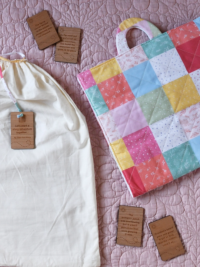 Several wooden story cards lay on a pink quilt beside a white cotton bag and a patchwork quilted advent calendar.
