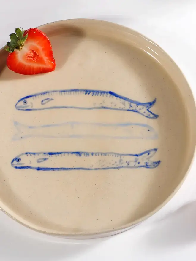 Handmade Ceramic Side Plate With 3 Blue Sardines Design, a side plate with a fish design sitting on a white table surface, accompanied by a raspberry. 