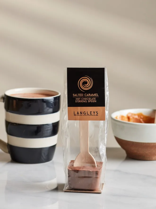 Milk Hot Chocolate Salted Caramel Stirring Spoon, a chocolate serving spoon pictured on a marble table top next to a hot chocolate and a bowl of food. 