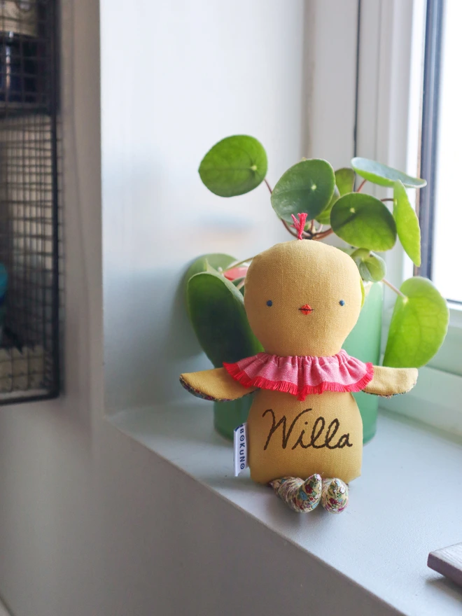 Handmade chick doll made from soft yellow fabric, with a stitched face, small beak, ruffled collar, and patterned fabric arms and long legs.