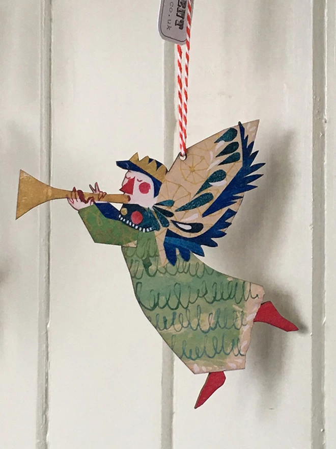 Joyful Angel illustrated wooden Christmas decorations in green, gold and red. The decoration hangs against a green panelled wall.