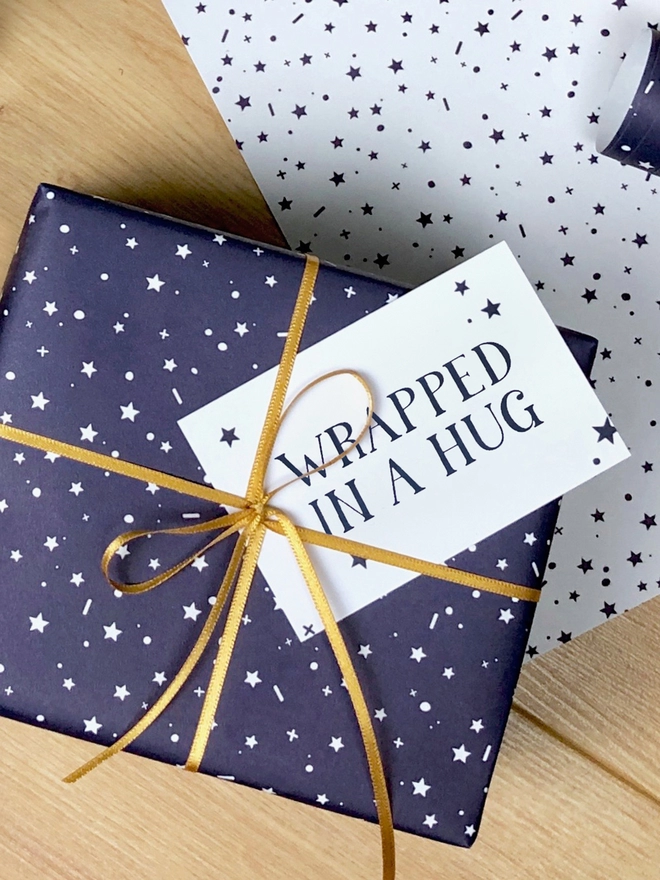 A gift wrapped in navy blue wrapping paper with a tiny star design and a gift tag that reads "Wrapped in a hug" is on a wooden surface.