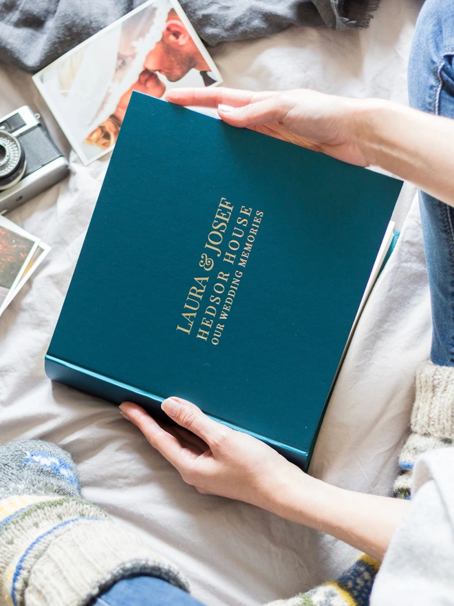 a woman on a bed is holding a blue wedding guest book in her hands. The wedding guest book has been printed on the front with gold lettering