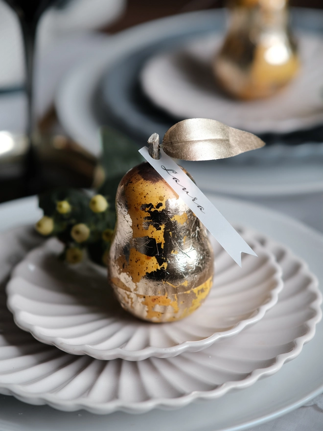 Gilded vintage-style Christmas pear ornament opened to reveal a small surprise inside, shown on a festive Christmas table setting with white plates and ribbon.