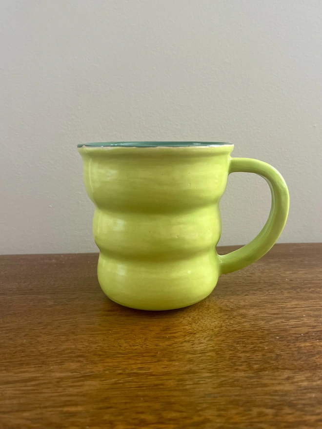 Hand-thrown 400 ml ceramic coffee mug with a wavy shape, bright lime green glaze, forest green interior and rounded handle, handmade by Hannah Inskip Pottery and sitting on a wooden table