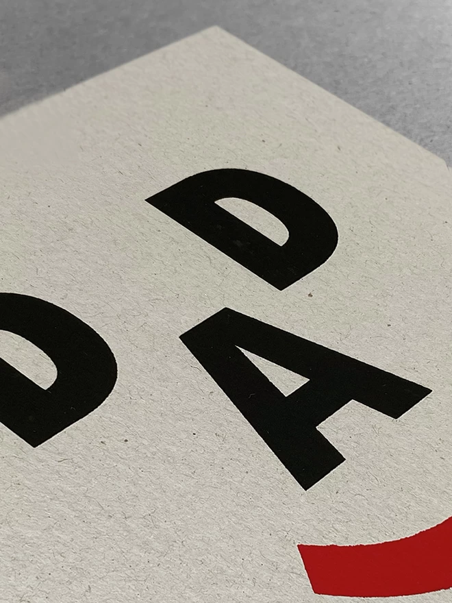 Close up of a greetings card, the letters D, A, D and a closed bracket symbol make up a happy face, screenprinted on recycled grey card. 