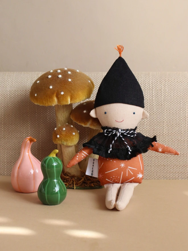 Handmade autumn elf doll with a black pointed hat, orange patterned top, black ruffled collar with black-and-white twine tie, embroidered blue eyes and pink smile, and light coloured face and legs 