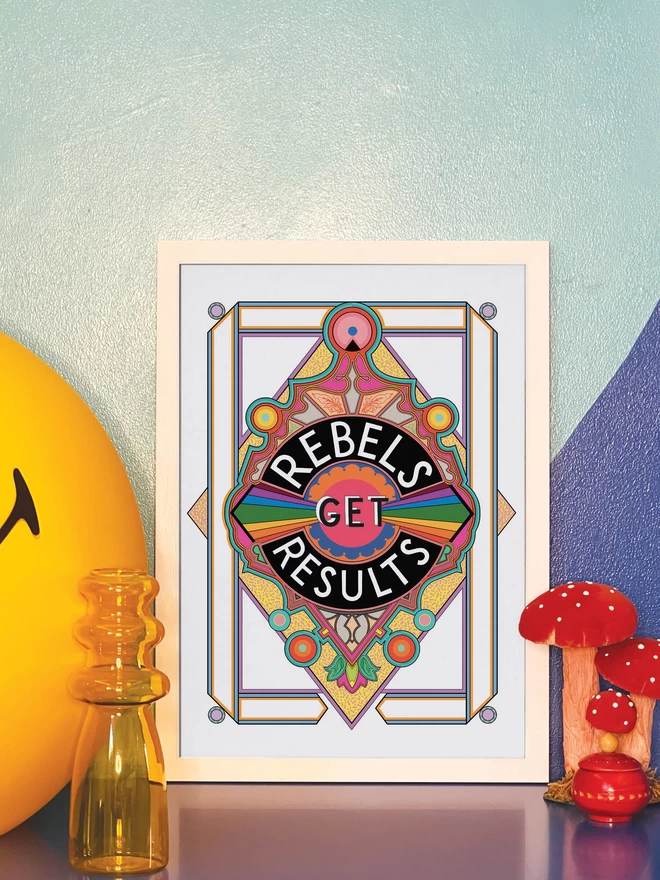 Rebels Get Results is written in white on a black background at the centre of this vibrant, abstract portrait illustration, with a white background and rainbows emitting from the centre, and multi-coloured detailing. The picture is in a white frame leaning against a painted blue wall on a blue cabinet. Next to the frame is a large illuminated yellow Smiley lamp, a yellow vase, a model of three red and white toadstools and a small round red wooden pot.