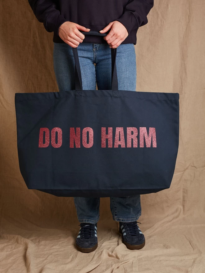 Model holding a massive navy cotton canvas tote bag with the words Do No Harm printed in pink glitter