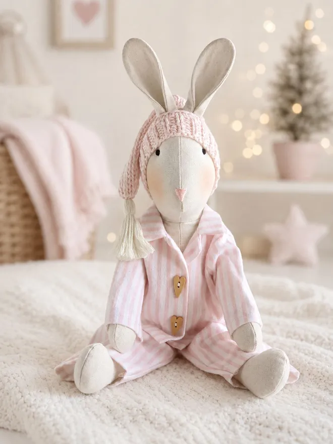 Christmas Eve Veronica Bunny wearing pink striped pyjamas and a knitted sleepy hat in a pale Christmas nursery with warm twinkle lights.