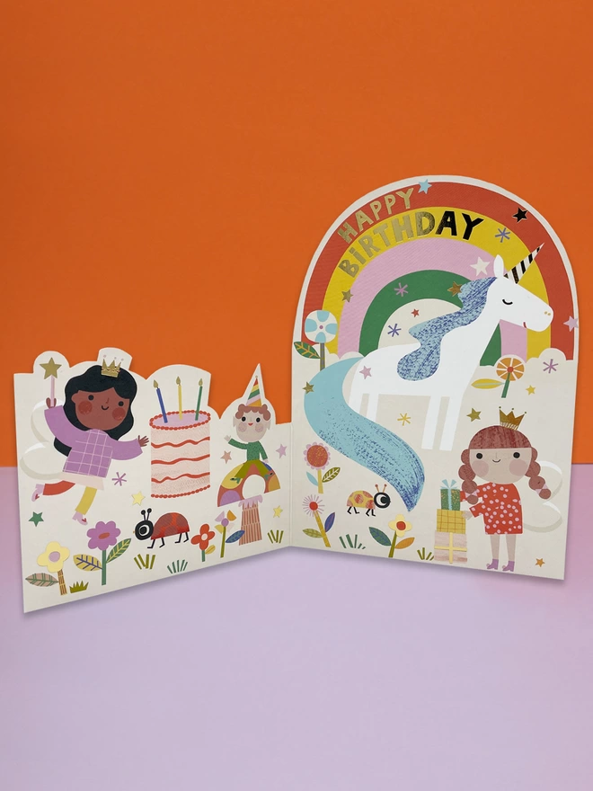 A magical fairy and unicorn themed, die cut children’s birthday card, with a colourful rainbow, magical stars, flowers and a giant birthday cake. The card is complete with gold foil details and a gold foil ‘Happy Birthday’ message